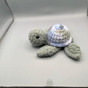 May include: A handmade crochet turtle in shades of green, blue, and gray. The turtle has a textured shell with a blue and gray pattern, a round head, and four flipper-like feet. The turtle is a soft, plush toy.