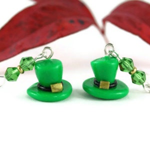 Puede incluir: Un par de pendientes de sombrero de duende verde con detalles de cristal verde. Los pendientes están hechos de arcilla polimérica y tienen ganchos de plata.
