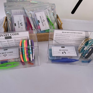 May include: Two clear plastic containers filled with beading kits. The container on the left has a chart for a binary code and a secret code. The container on the right has a sheet of instructions for beading kits and a variety of blue and white beaded bracelets.