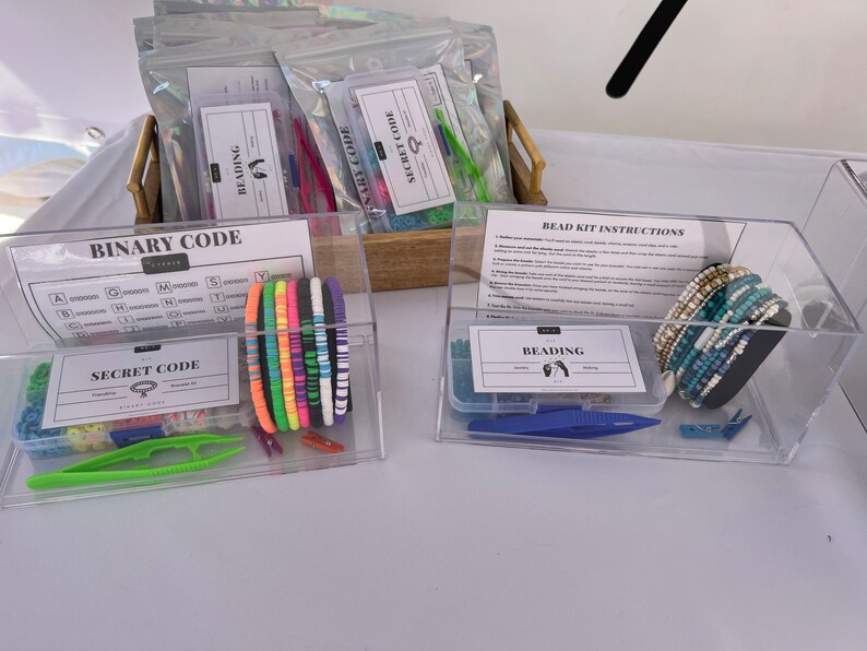 May include: Two clear plastic containers filled with beading kits. The container on the left has a chart for a binary code and a secret code. The container on the right has a sheet of instructions for beading kits and a variety of blue and white beaded bracelets.