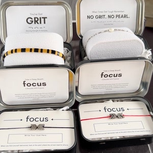 May include: Four small metal tins with lids, each containing a bracelet and a motivational message. The tins are labeled with the words "Grit", "No Grit, No Pearl", "Focus", and "Focus". The bracelets are black and gold, silver, red and silver, and black and silver.