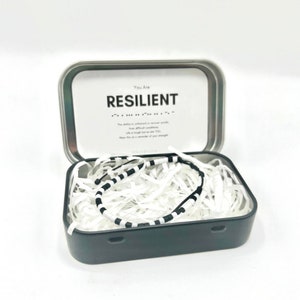 May include: A black and white striped bracelet in a small tin box with a white label that reads "You Are RESILIENT". The ability to withstand or recover quickly from difficult conditions. Life is tough but so are YOU. Wear this as a reminder of your strength.