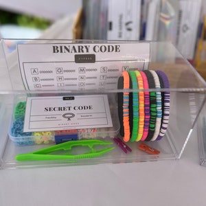 May include: A clear plastic container with a variety of colorful bracelets, beads, and tools for making friendship bracelets. The container also includes a card with the text "Secret Code Friendship Bracelet Kit" and a chart with the text "Binary Code" and a list of letters and their corresponding binary code.