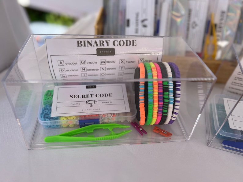 May include: A clear plastic container with a variety of colorful bracelets, beads, and tools for making friendship bracelets. The container also includes a card with the text "Secret Code Friendship Bracelet Kit" and a chart with the text "Binary Code" and a list of letters and their corresponding binary code.