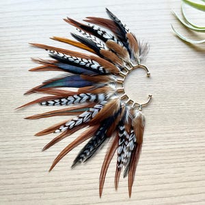 Può includere: Un ear cuff con una montatura in metallo dorato ornata da una varietà di piume. Le piume sono nelle tonalità del marrone, nero, bianco e verde acqua, creando un effetto stratificato e strutturato. L'ear cuff è progettato per avvolgere l'orecchio.