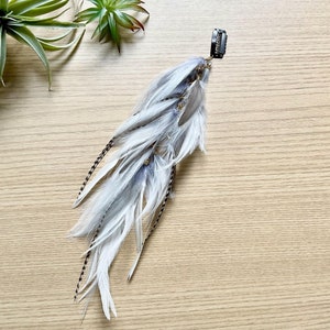 May include: A long, white feather hair clip with black accents. The clip is attached to a bunch of white feathers with a few black feathers mixed in.