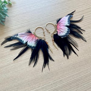 May include: A pair of ear cuffs featuring butterfly wing designs. The wings are pink, white, and black, with black feathers extending from the base. The ear cuffs have a gold-colored hoop and are adorned with small beads and charms.