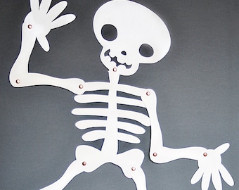 May include: A white, articulated skeleton decoration with copper rivets, posed against a gray background. The skeleton is dancing, with a jack-o'-lantern and several white pumpkins in the foreground, and a teal velvet pumpkin.