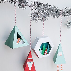 May include: A collection of geometric Christmas ornaments hanging from a silver garland. The ornaments are various shapes, including triangles and a hexagon, and feature festive designs like Santa Claus, a fox, and a winter forest scene. Each ornament is suspended by a red and white string.