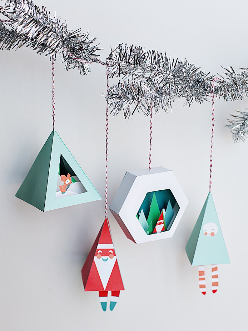 May include: A collection of geometric Christmas ornaments hanging from a silver garland. The ornaments are various shapes, including triangles and a hexagon, and feature festive designs like Santa Claus, a fox, and a winter forest scene. Each ornament is suspended by a red and white string.