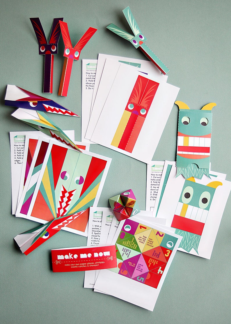 May include: A collection of colorful paper craft kits, including paper airplanes, monsters, and a geometric shape. The kits feature red, green, and yellow accents, with instructions and the text "make me now".