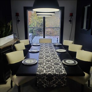 May include: A formal dining table set for a meal. The black table is set with white plates with a blue floral design, a black and white patterned table runner, and cream-colored chairs with black pillows.