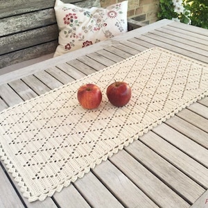 Op de afbeelding: Een gehaakte tafelloper met een ruitpatroon in crèmekleur. De loper ligt op een houten tafel met twee rode appels erop.