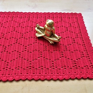 Può includere: Un runner da tavolo in crochet rosso con un bordo a festoni. Una statuetta di rana dorata è seduta sul runner.