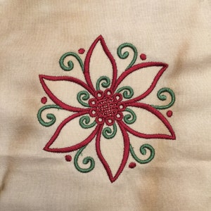 May include: A red and green embroidered flower design on a beige fabric background.
