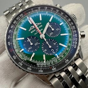 May include: A stainless steel Breitling Navitimer watch with a green and blue dial. The watch features three sub-dials, a date display, and a rotating bezel with various markings. The watch band is made of stainless steel.