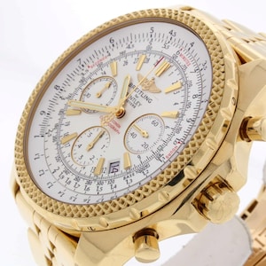 Könnte beinhalten: Eine goldfarbene Breitling Bentley Motors Chronographenuhr. Die Uhr hat ein weißes Zifferblatt mit mehreren Hilfszifferblättern, goldenen Zeigern und einer Datumsanzeige. Die Lünette und das Armband sind ebenfalls goldfarben, mit einem strukturierten Design auf der Lünette.