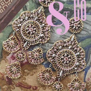 Może przedstawiać: Para kolczyków z koralików w kolorze różowym i złotym. Kolczyki są wykonane z małych koralików i mają geometryczny wzór. Kolczyki są pokazane na tle wzorzystej tkaniny. W obrazie widoczny jest tekst "ORECCHINI IN BRICK STITCH. ALESSANDRA DEL VITTO PER ADELSLABORATORY".