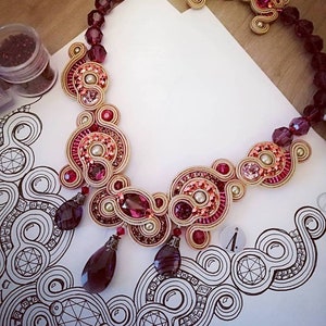 May include: An ornate necklace featuring a complex design with burgundy, pink, and gold accents. The necklace incorporates beads, crystals, and teardrop-shaped pendants. The jewelry is displayed on a white surface with a decorative black and white pattern.