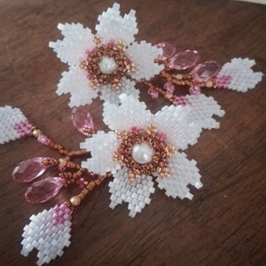 May include: A pair of white beaded earrings with pink and gold accents. Each earring features a flower design with a pearl in the center. The earrings are dangling and have a beaded chain with pink and gold beads.