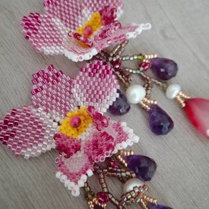 May include: Close-up of two beaded flower earrings. The flowers are pink, white, and yellow, with dangling amethyst, pearl, and pink teardrop-shaped beads. The earrings are handmade and feature intricate beadwork.