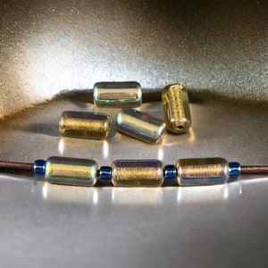 May include: A close-up of six iridescent gold glass beads. The beads are cylindrical and have a textured surface. The beads are arranged in a line on a gold surface.
