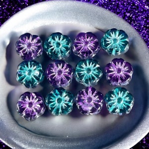 May include: Twelve purple and teal glass flower beads with a starburst design. The beads are arranged in a circle on a white plate.