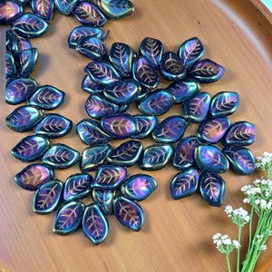May include: A collection of iridescent leaf-shaped beads in shades of blue, purple, and gold. The beads have a metallic sheen and a detailed leaf vein pattern. They are scattered on a wooden surface, suggesting a craft or jewelry-making theme.