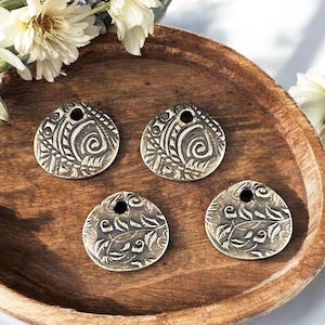 May include: Four antique brass-colored pendants with intricate embossed designs of swirling patterns and leaves. Each pendant has a small hole at the top for stringing. The pendants are displayed on a wooden tray, with white flowers in the background.