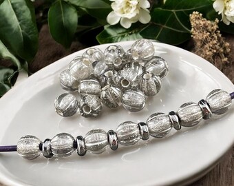 May include: A collection of clear, textured glass beads, some loose and some strung on a purple cord. The beads have a silver-like appearance and are displayed on a white plate. The beads are round with a ribbed design.