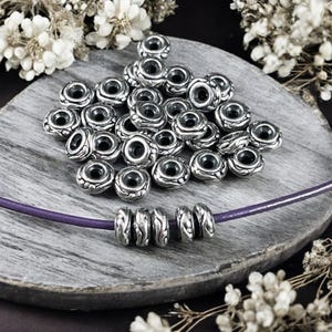 May include: A pile of silver metal beads with a decorative design. The beads are arranged on a wooden surface with a purple cord and more beads strung on it.