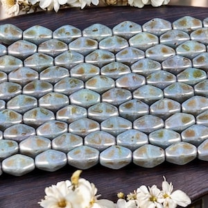 Pode incluir: Close-up de uma cole&ccedil;&atilde;o de contas hexagonais iridescentes. As contas t&ecirc;m um brilho perolado com toques de azul e dourado. As contas est&atilde;o dispostas em fileiras ordenadas, sugerindo que s&atilde;o para fazer joias ou artesanato.