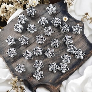 May include: A collection of silver-toned metal flower-shaped beads. The beads are arranged on a wooden surface.