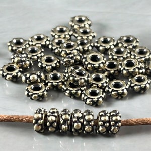 May include: A pile of gold-toned metal beads with a textured surface. The beads are round with a hole in the centre. A strand of the beads is strung on a brown leather cord.