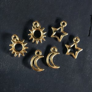 May include: Six gold-colored metal charms in the shape of a sun, star, crescent moon, and two stars. The charms are on a dark gray surface.