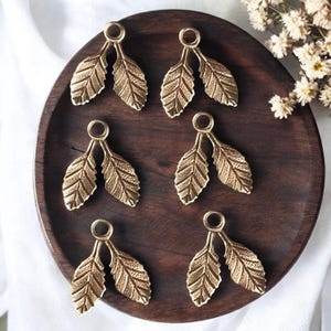 May include: Six gold leaf-shaped metal charms with a loop at the top. The charms are arranged in two rows of three on a wooden surface.