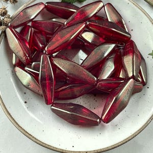 May include: A collection of elongated, red glass beads with a metallic sheen, displayed on a white ceramic plate. The beads have a faceted shape, catching the light and creating highlights. The beads are likely for jewelry making or crafting.
