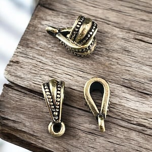 May include: Three antique brass-colored jewelry findings on a wooden surface. The findings include a bail, a connector, and a decorative clasp. The bail has a loop for attaching to a chain or other jewelry component.