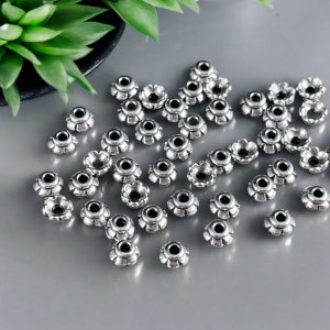 May include: A collection of small, silver-colored metal flower-shaped beads. Each bead has a central hole and petal-like edges. The beads are scattered on a gray surface, with a green plant visible in the background.