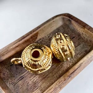 May include: Two gold-toned metal beads with a polished finish. One bead is round and features a central hole with radiating lines, while the other is barrel-shaped with vertical ridges and a series of small raised dots around its center.