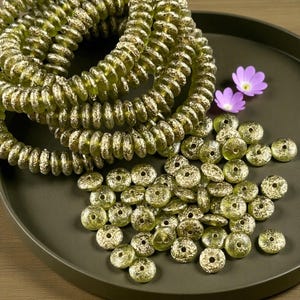May include: Olive green glass beads and coiled strands on a dark green tray. The beads are round with a textured, metallic finish. Two strands of coiled beads are in the background. Two small purple flowers are also present.