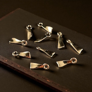 May include: A collection of small, gold-colored metal clasps, each with a unique shape and a small loop at one end. The clasps are scattered on a dark brown surface, creating a visually appealing arrangement. These clasps are likely used for jewelry or other craft projects.