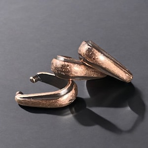 May include: Three bronze-colored hoop earrings with a hammered texture are displayed against a dark background. The earrings have a curved design with a small post and a rounded, textured body. The light catches the metal, highlighting the details.