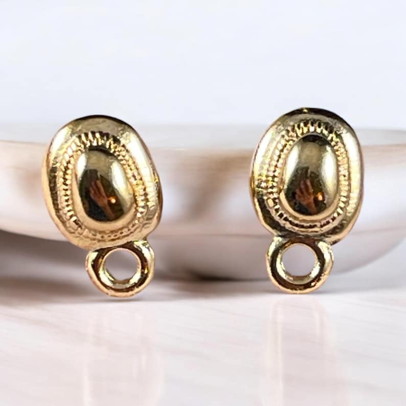 May include: Two gold-colored earring findings. Each finding has an oval shape with a textured surface and a small loop at the bottom. The findings are displayed on a white surface.