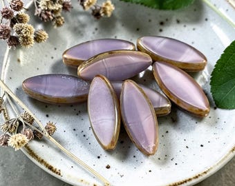 May include: Oval-shaped lavender beads with a golden rim are displayed on a speckled ceramic plate. The beads are arranged in a cluster, showcasing their smooth, polished surface. Dried floral accents and a green leaf add a natural touch to the composition.