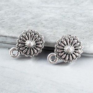 May include: Two silver-colored, daisy-shaped earring components. Each component has a detailed floral design with a central raised circle and a small loop for attachment. The earrings are set against a light gray background.
