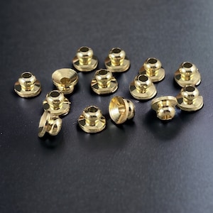 May include: A collection of small, shiny brass rivets with a rounded top and a flat base. The rivets are scattered on a dark surface, showcasing their metallic luster and detailed design. These fasteners are likely used in leathercraft or similar projects.