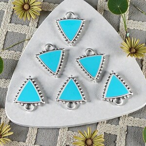 Peut inclure: Six breloques triangulaires turquoise avec des bords perlés et des boucles argentés. Les breloques sont disposées sur une surface triangulaire grise, avec des fleurs jaunes et de la verdure en arrière-plan. Idéal pour la création de bijoux.