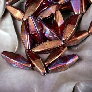 May include: Close-up of a collection of iridescent beads. The beads are elongated, with a faceted surface that reflects shades of bronze, purple, and gold. They are strung together, suggesting a necklace or bracelet. The background is a soft, neutral fabric.