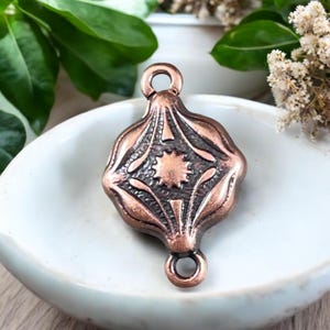 May include: A copper-colored pendant with an ornate, symmetrical design. The pendant features a central starburst pattern and decorative lines. It has a loop at the top and bottom for attaching to a chain or other jewelry component.